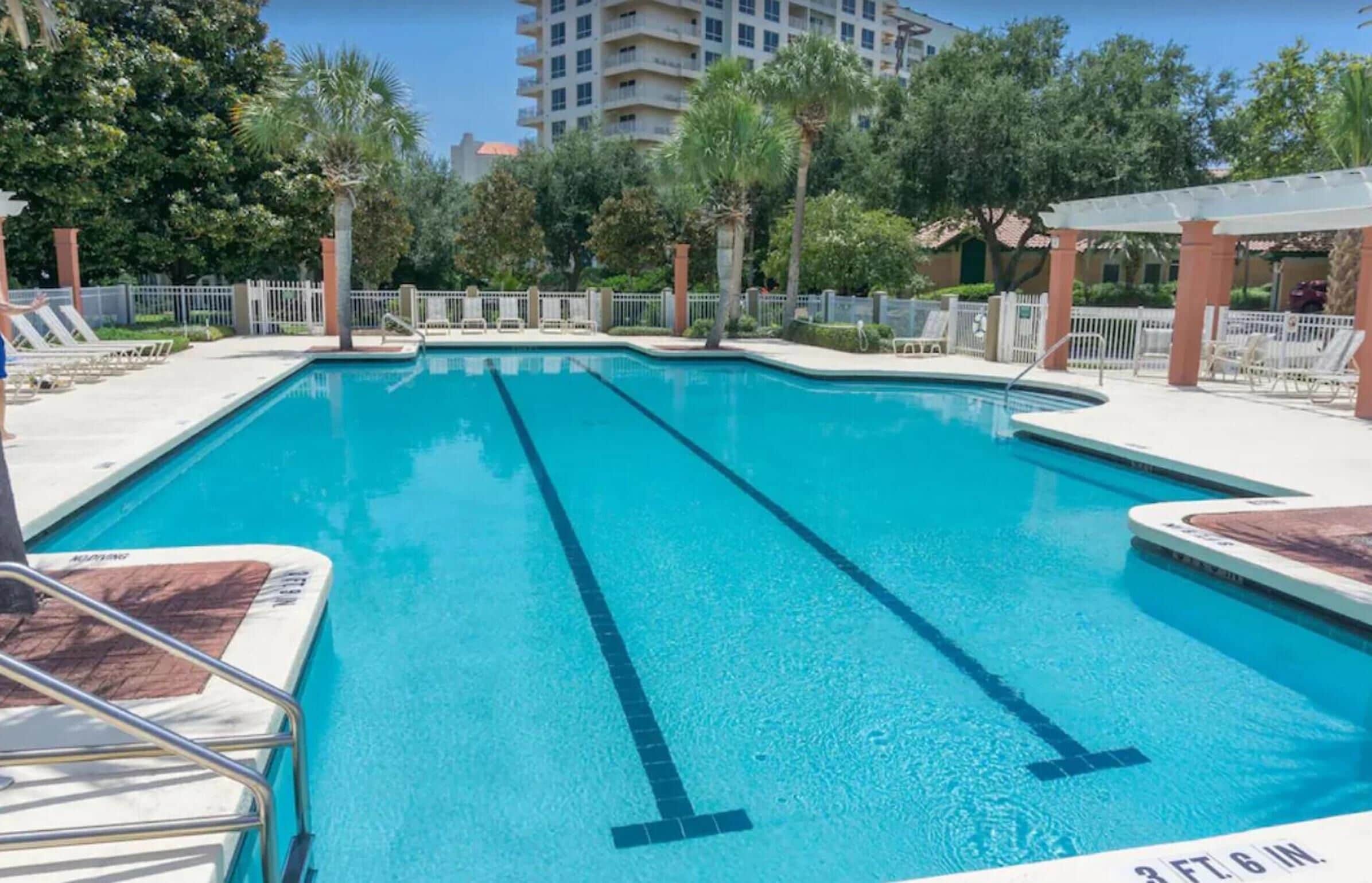 A clear blue pool with lane dividers, surrounded by lounge chairs and a white fence.