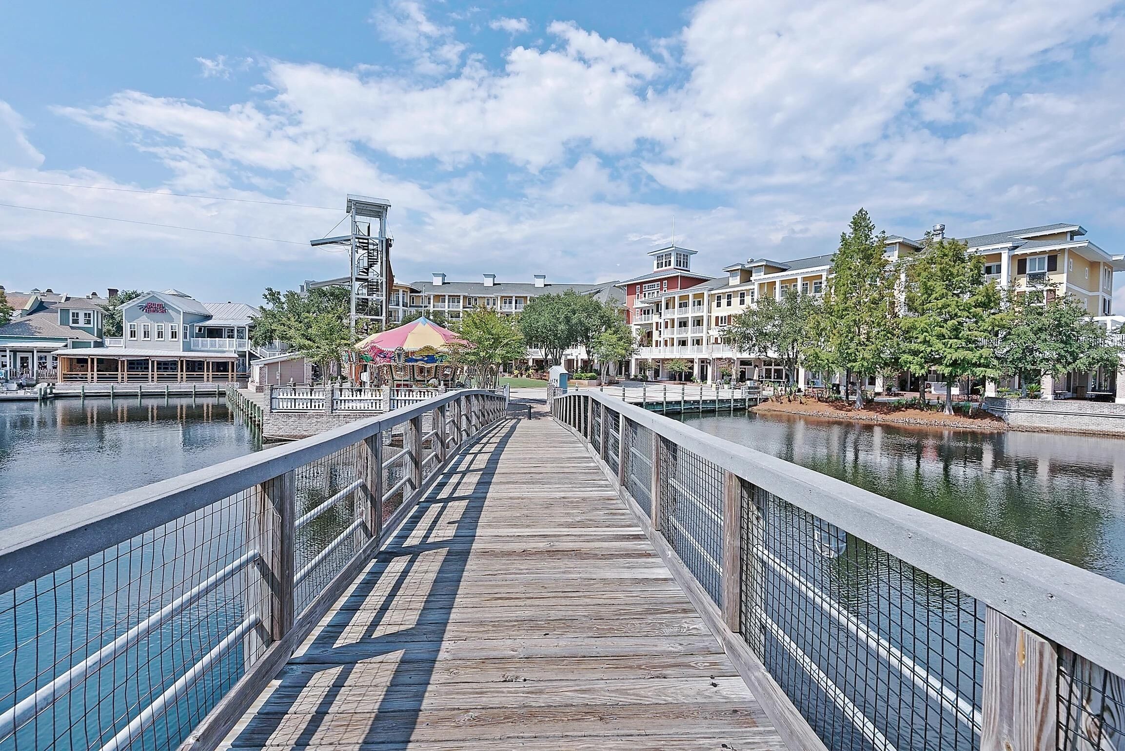 A wooden boardwalk with a metal railing, leading to a waterfront area with buildings and trees.