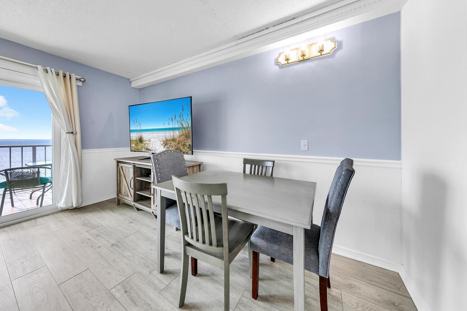 A dining area with a table and chairs, a cabinet, a painting, and a balcony with a view of the beach.