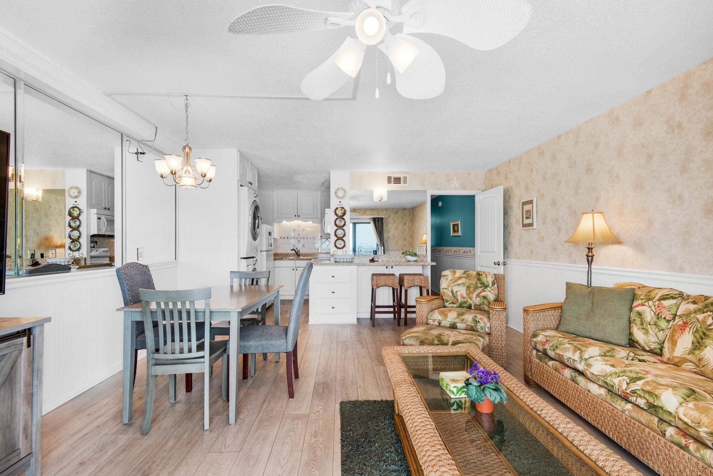 A spacious living area with a dining table, chairs, a sofa, and a coffee table. There is a ceiling fan, a chandelier, and a lamp.