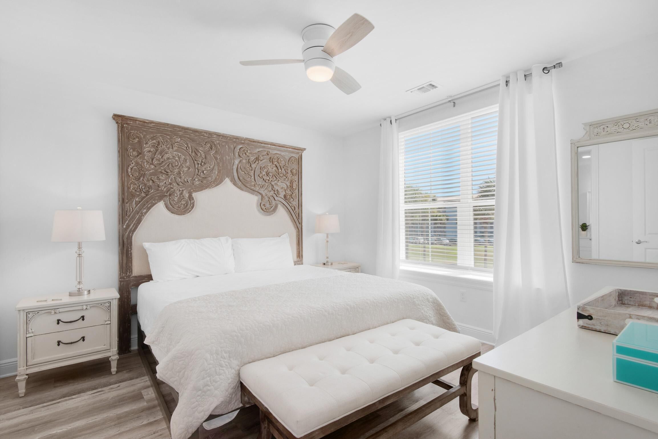 Une chambre à coucher avec un grand lit, une tête de lit en bois, un banc, une table de chevet, une lampe, un ventilateur de plafond, une fenêtre avec des rideaux et un miroir.