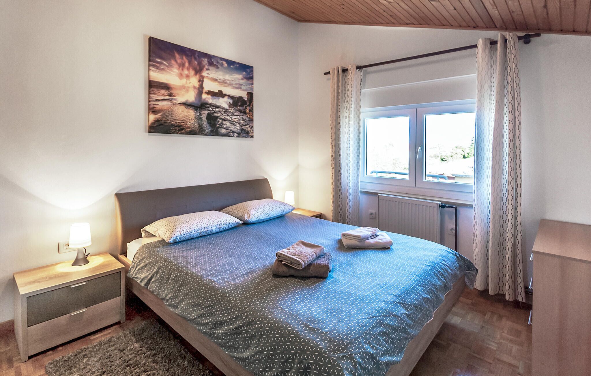 A bedroom with a bed, a nightstand, a lamp, a window with curtains, and a painting on the wall.