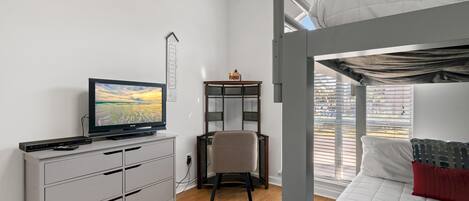 Un dormitorio con una cama individual superior e inferior, un escritorio con silla, un televisor y una ventana con persianas.