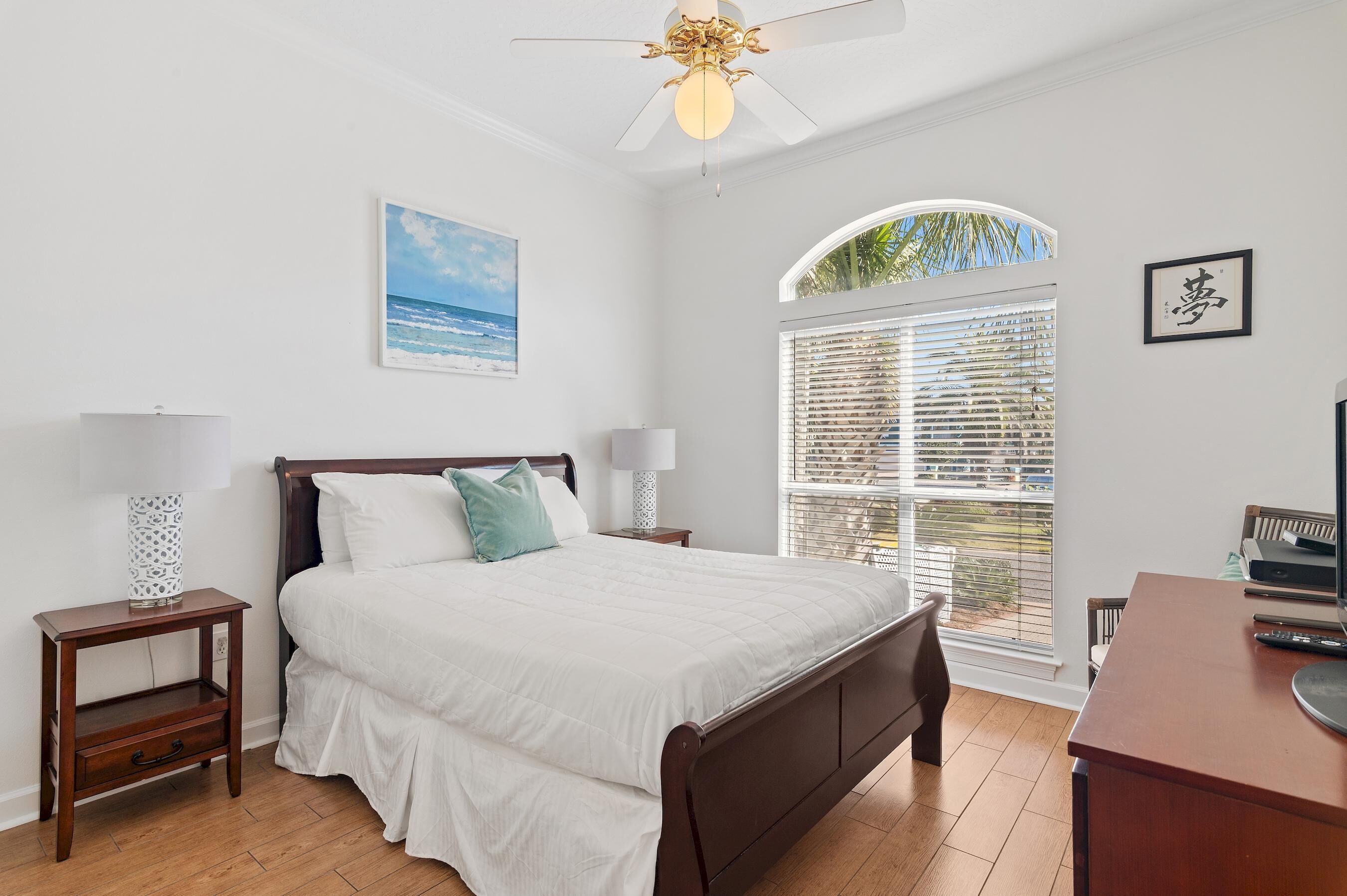 A bedroom with a bed, two bedside tables, a desk, a ceiling fan, a window with blinds, and a framed picture on the wall.