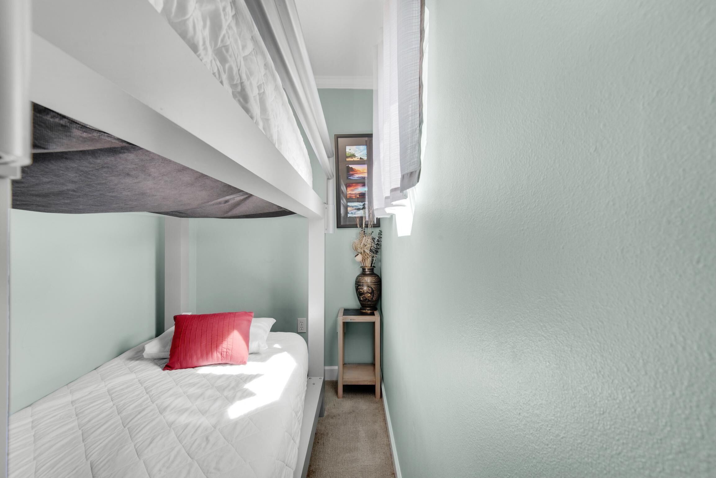 Een kleine kamer met een stapelbed, een klein nachtkastje met een vaas, en een ingelijst plaatje aan de muur.