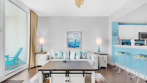 A modern living room with a sofa, coffee table, and two side tables with lamps. There is a kitchen area with white cabinets and a blue accent wall. - Majestic Beach Towers 1-2113 (Panama City Beach)