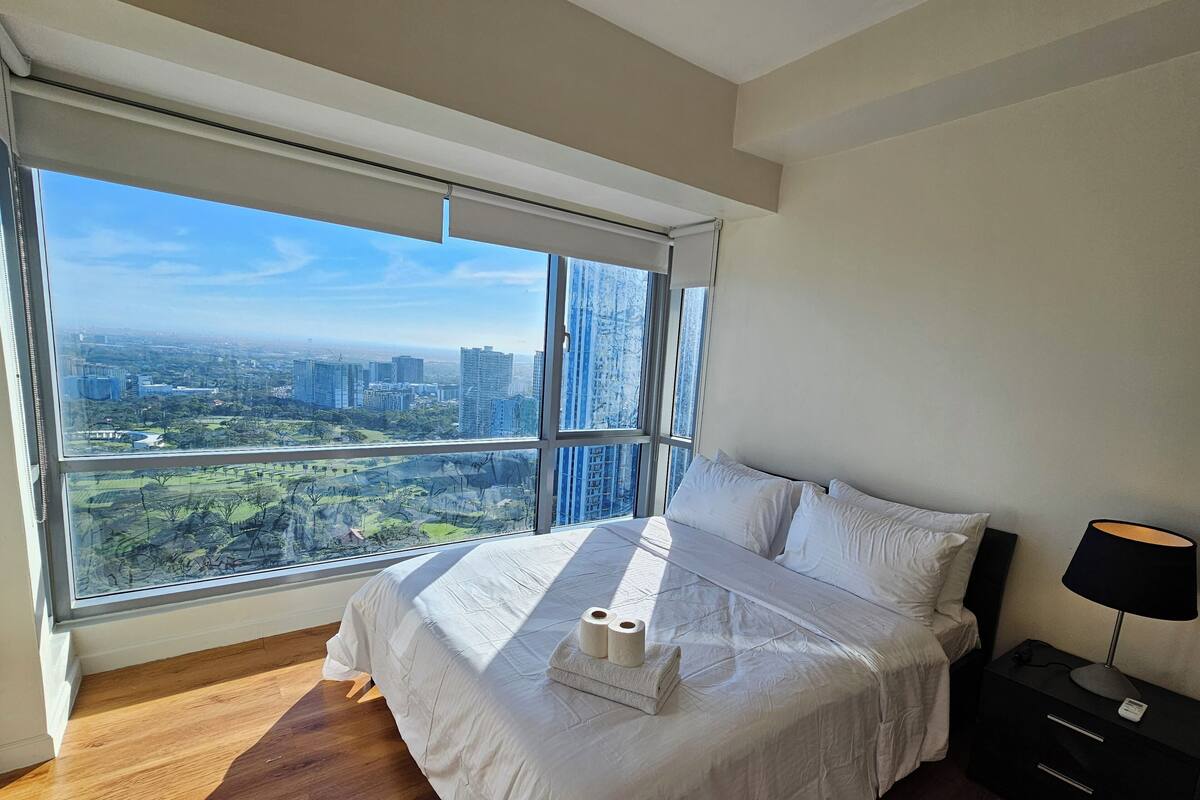 Un dormitorio con una ventana grande, una cama, una mesita de noche y vistas a un paisaje urbano.