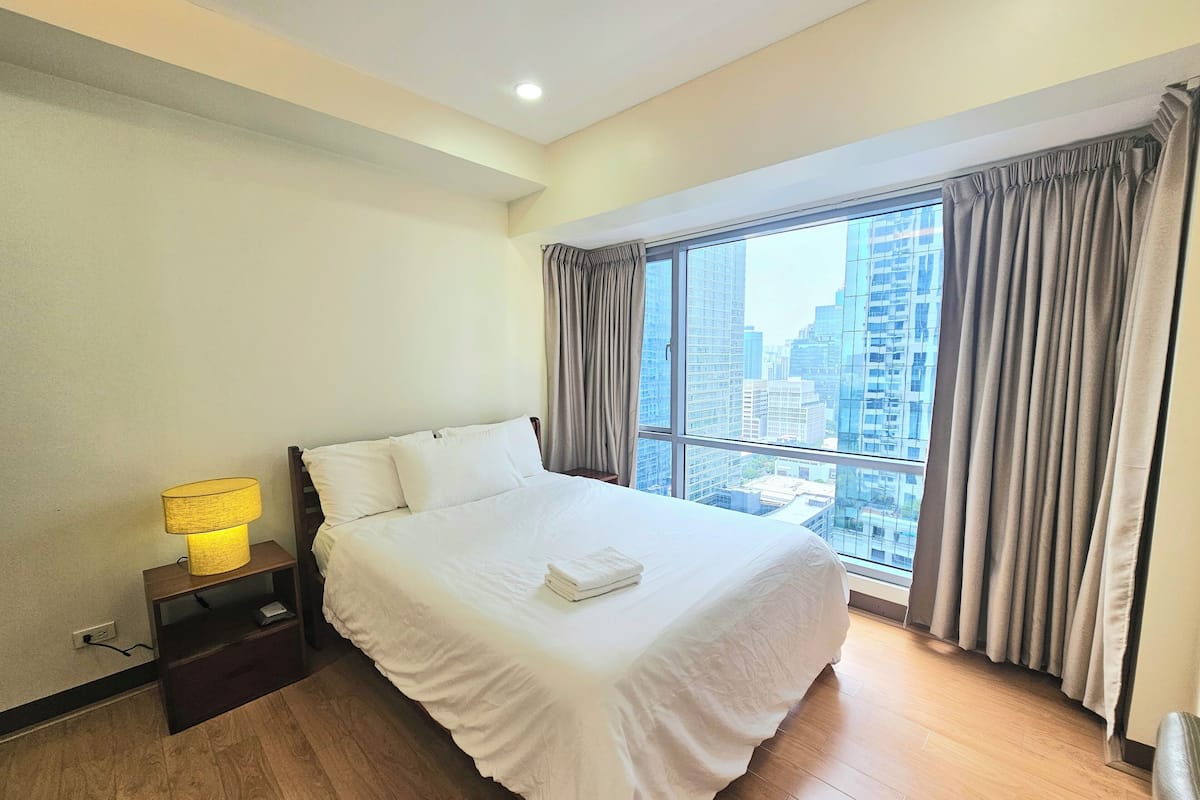 Habitación de hotel con una cama grande, una lámpara de noche y vistas de edificios altos a través del ventanal.