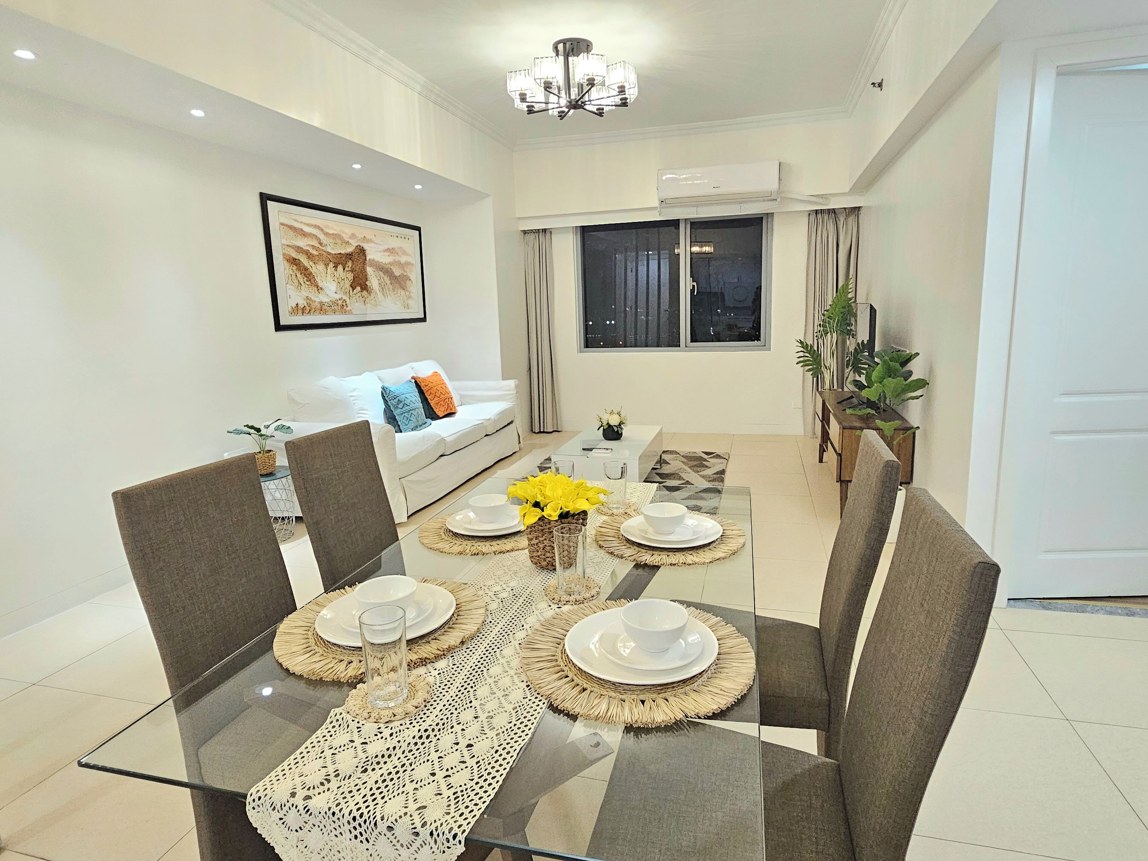 A modern dining area with a glass table, woven placemats, and a sofa in the background.