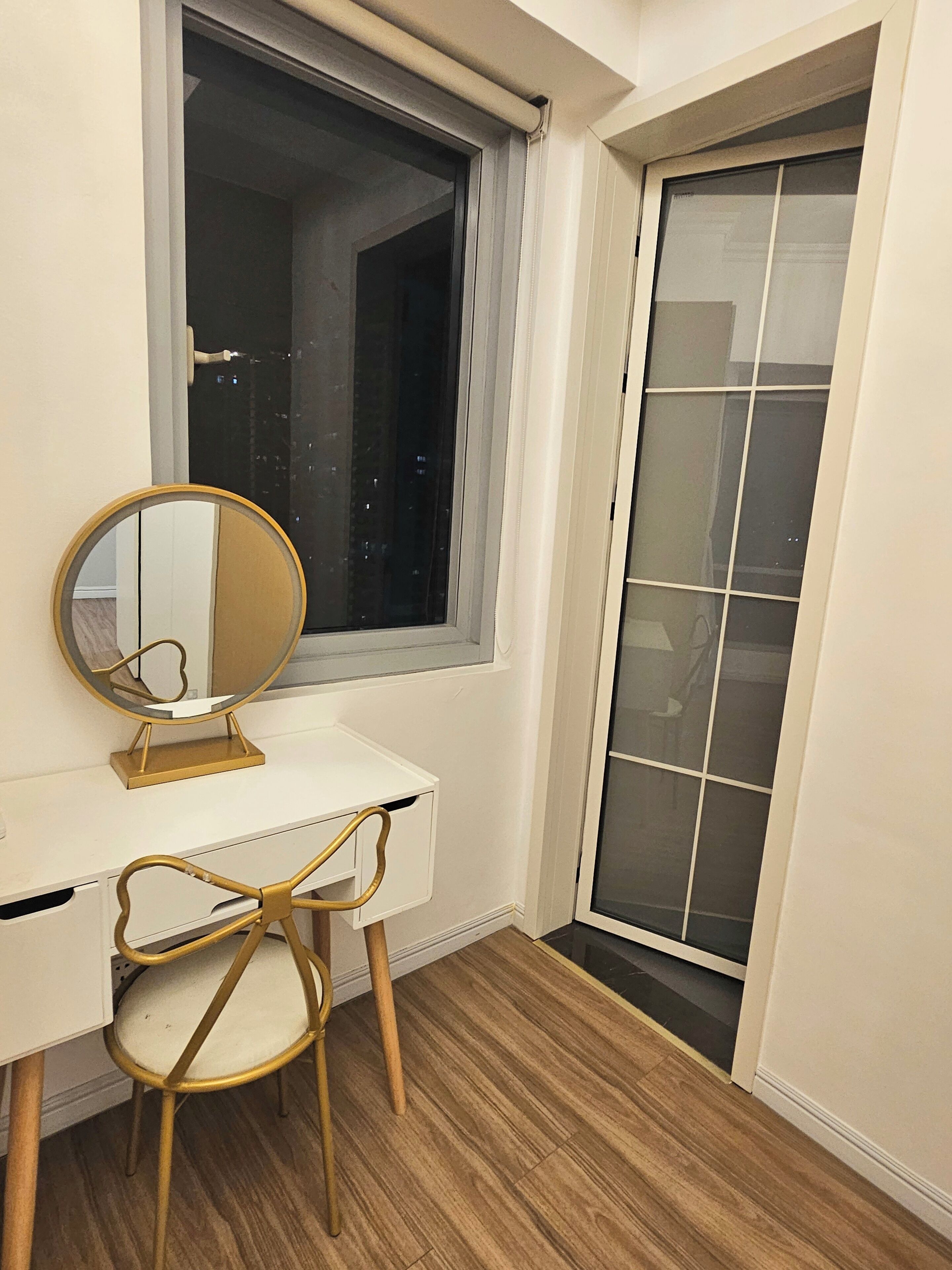 A white desk with a gold-framed mirror and a gold-framed chair in front of a window with a view of a cityscape.