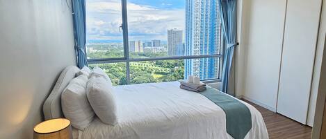 A hotel room with a large window, a bed with white linens, a lamp, and a view of a cityscape.