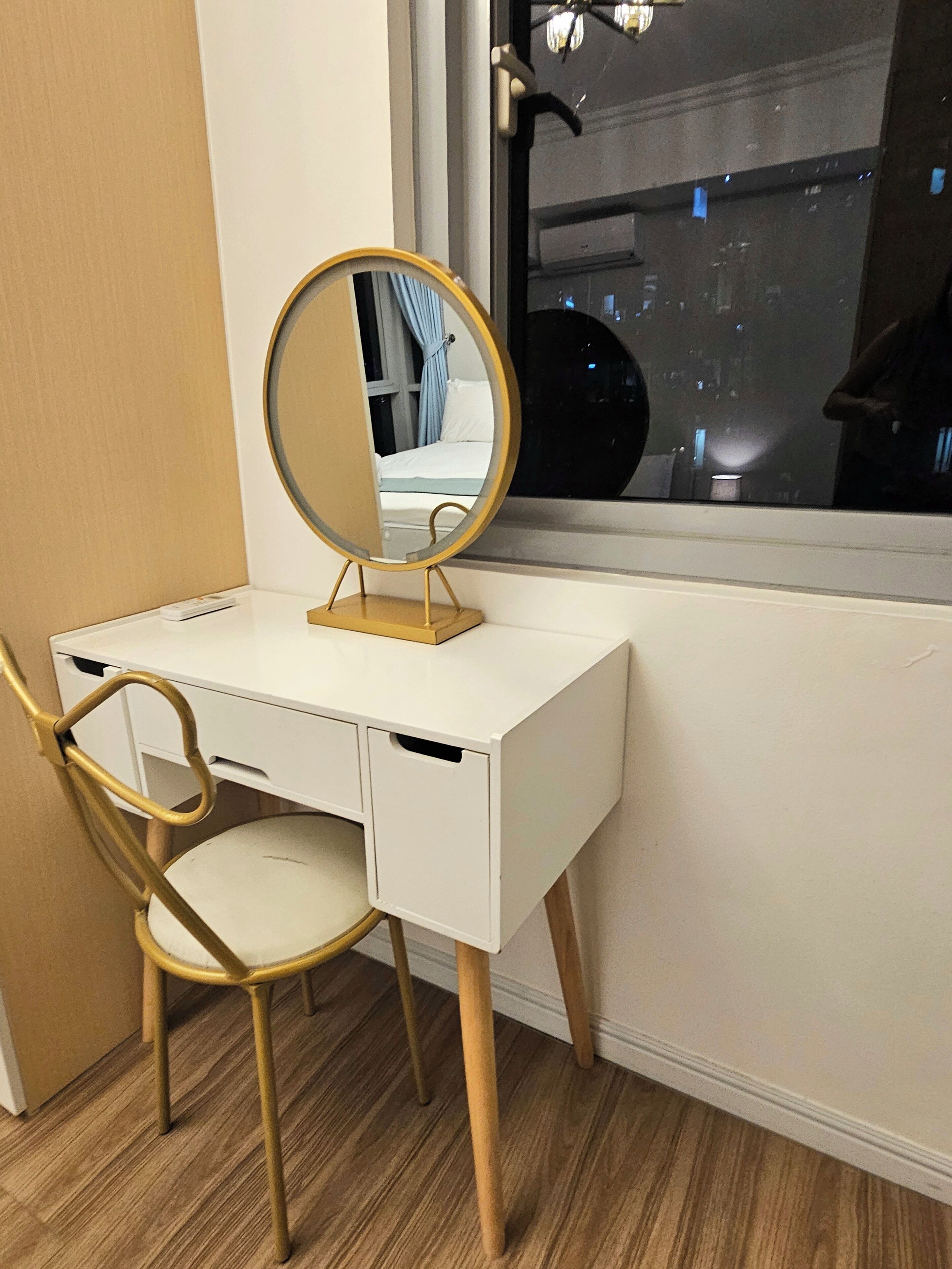 A white desk with a round mirror, a gold chair, and a window with a view of the city at night.
