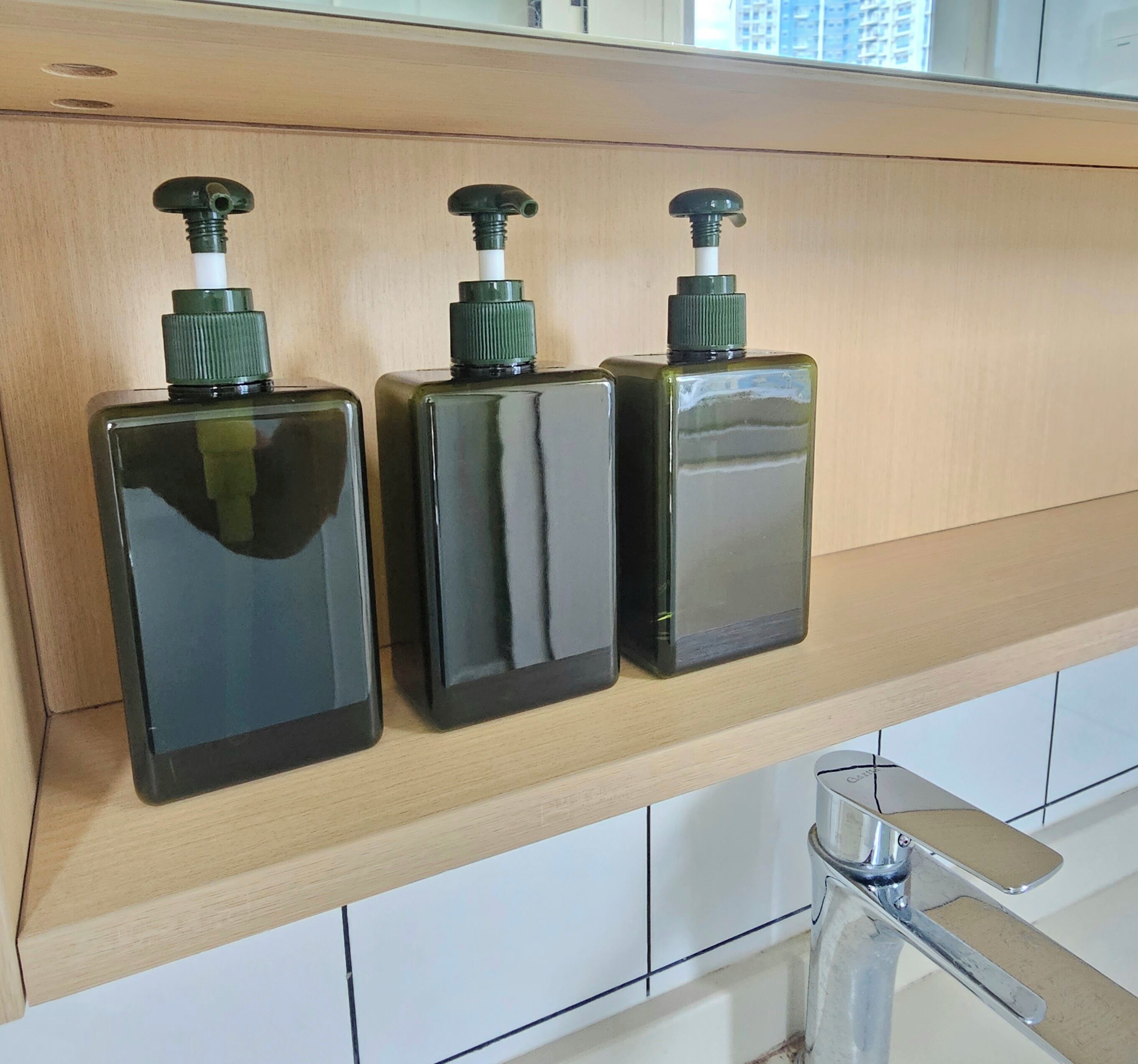 Three black bottles with green pumps on a wooden shelf in a kitchen.
