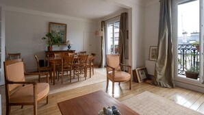 A living room with a wooden dining table, chairs, a sofa, and a large window with curtains. - Appartement Apalta - Welkeys (Paris)