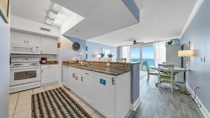 Eine moderne Küche mit weißen Schränken, Granitarbeitsplatten und einer großen Kochinsel. Es gibt einen Essbereich mit Tisch und Stühlen sowie einen Blick auf den Ozean durch große Fenster.