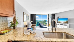 A modern kitchen with granite countertops, a sink, and a dishwasher. There is a dining area with a table and chairs, a potted plant, and a view of the ocean through large windows. - Edgewater Tower 1-305 (Panama City Beach)