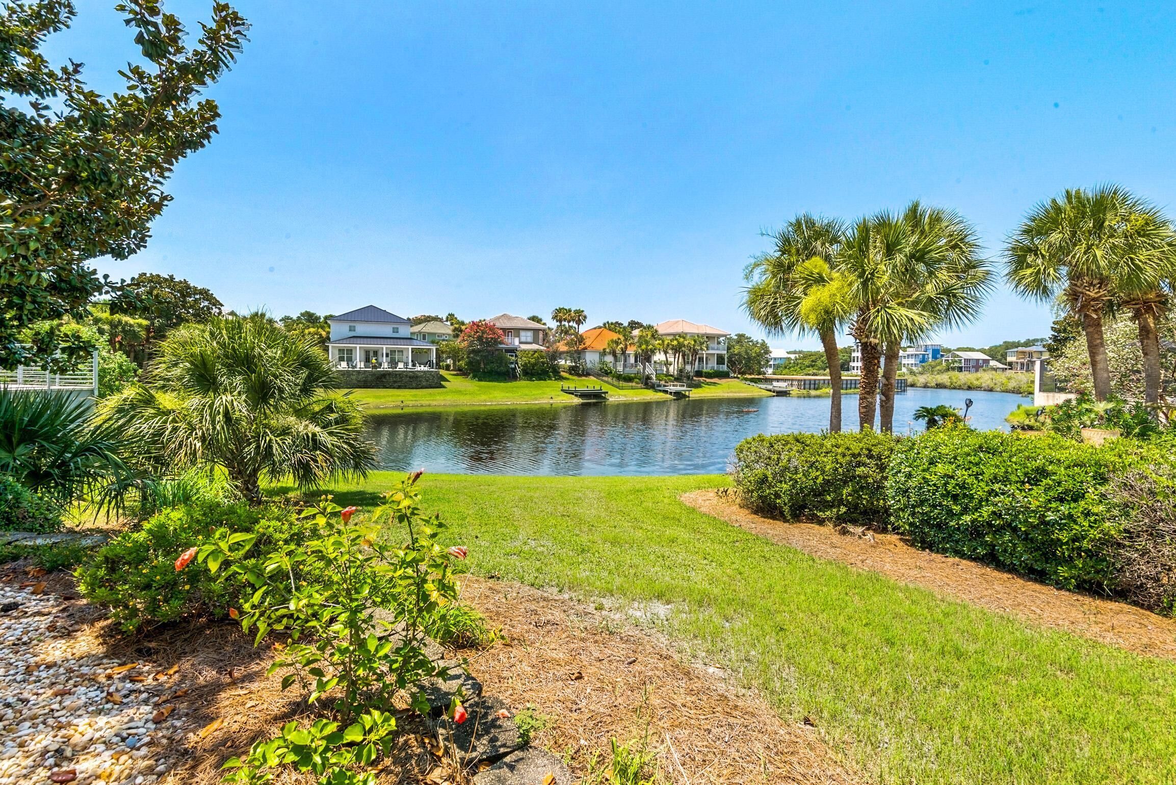 A lakeside view with a well-maintained lawn, palm trees, and a row of houses in the background.