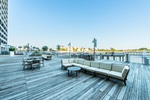 Eine Holzterrasse mit Sitzgelegenheiten im Freien, darunter eine Couch und ein runder Tisch, mit Blick auf einen Strand.