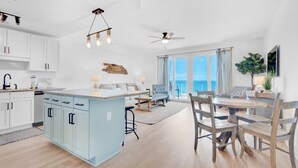 A modern kitchen with white cabinets, a light blue island, and pendant lighting. There is a dining area with a round table and chairs, and a living room with a sofa and a large window overlooking the ocean. - Laketown Wharf 1714 (Panama City Beach)