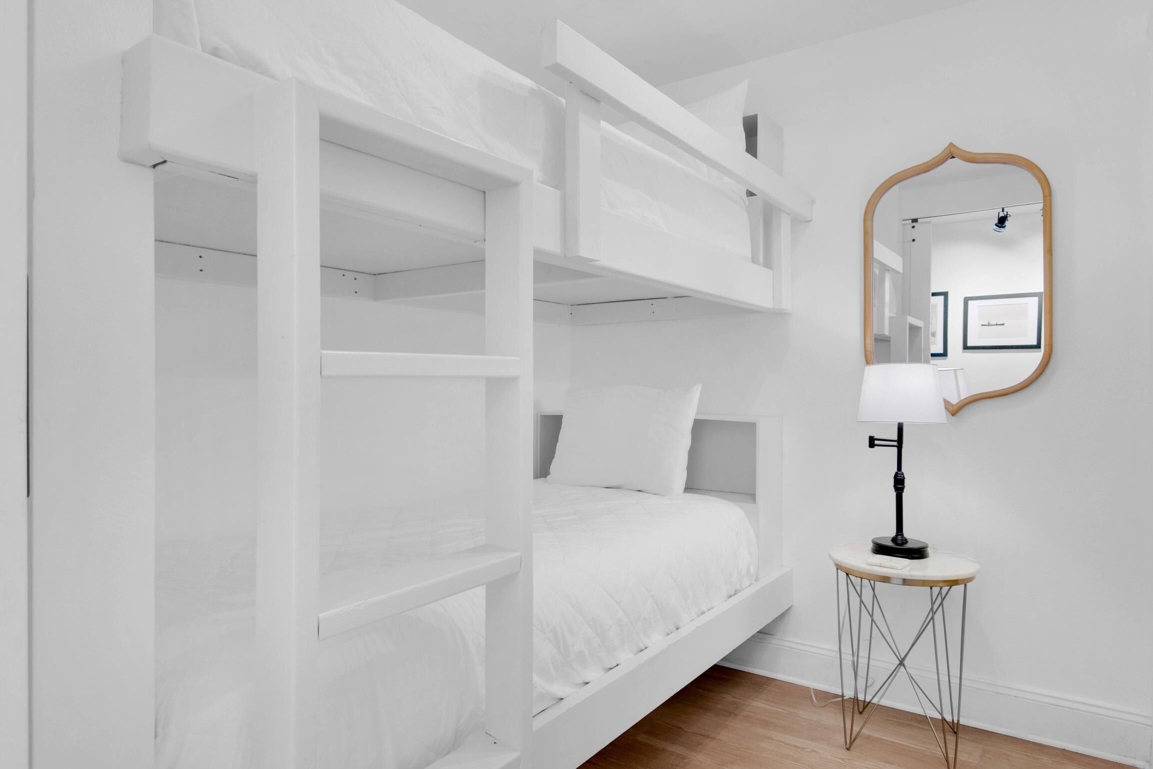 Een witte kamer met een stapelbed, een klein tafeltje met een lamp en een spiegel aan de muur.