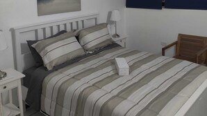 Quarto com cama, mesas de cabeceira, luminária, pintura e janela com cortinas.