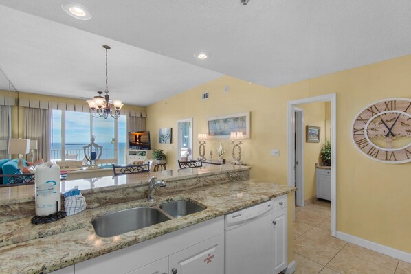 A kitchen with granite countertops, stainless steel sink, and white cabinets. There is a clock on the wall and a chandelier above the sink.