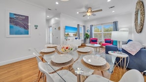 A modern living room with a dining area, a ceiling fan, and a large window with curtains. - Legacy 506- The Coastal Gem (Santa Rosa Beach)