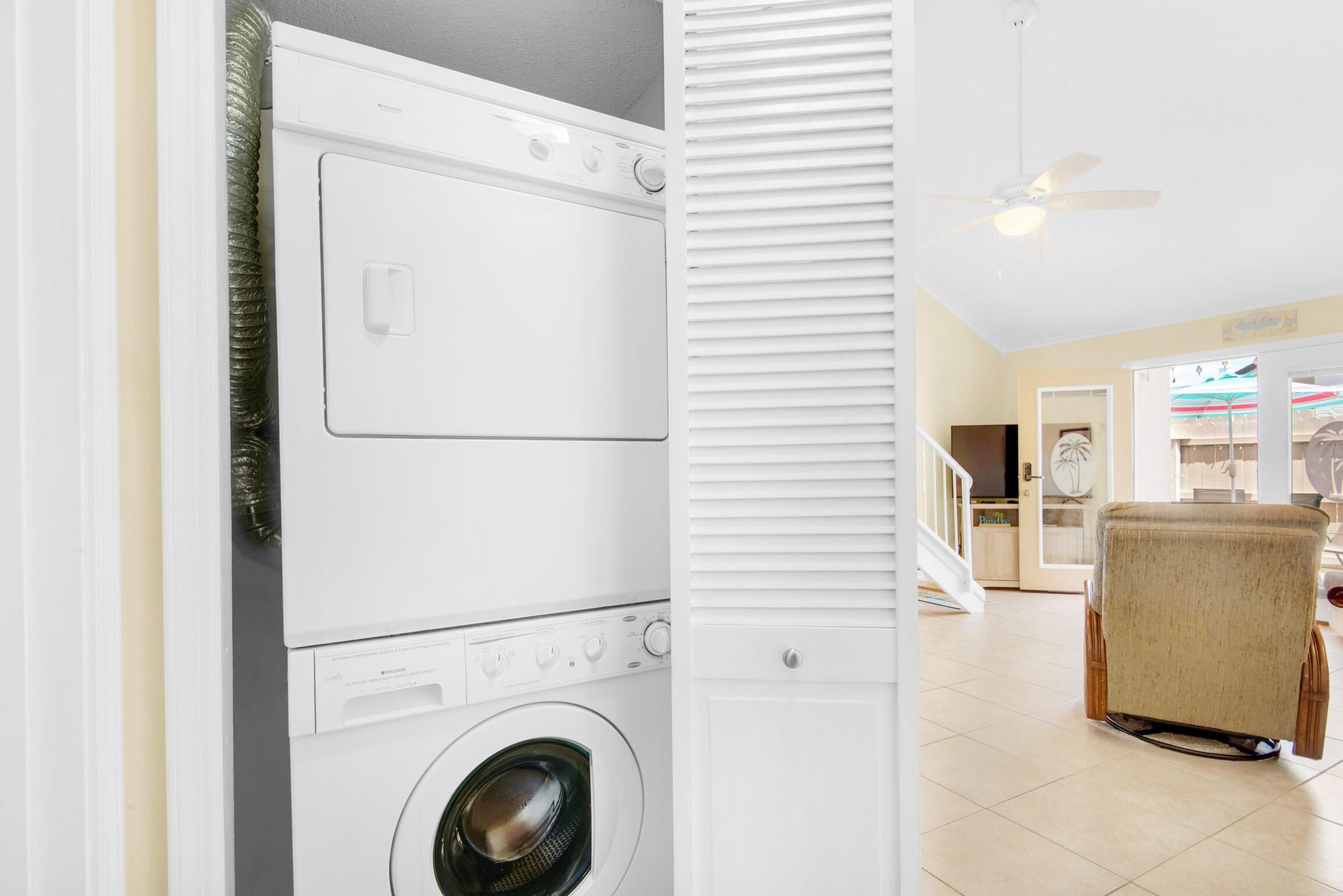 A laundry room with stacked washers and dryers, a ceiling fan, and a view of a living area with a chair and a door leading to another room.