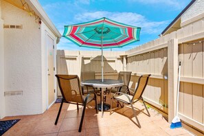 Een terras met een ronde tafel en stoelen, een kleurrijke parasol en een houten schutting.