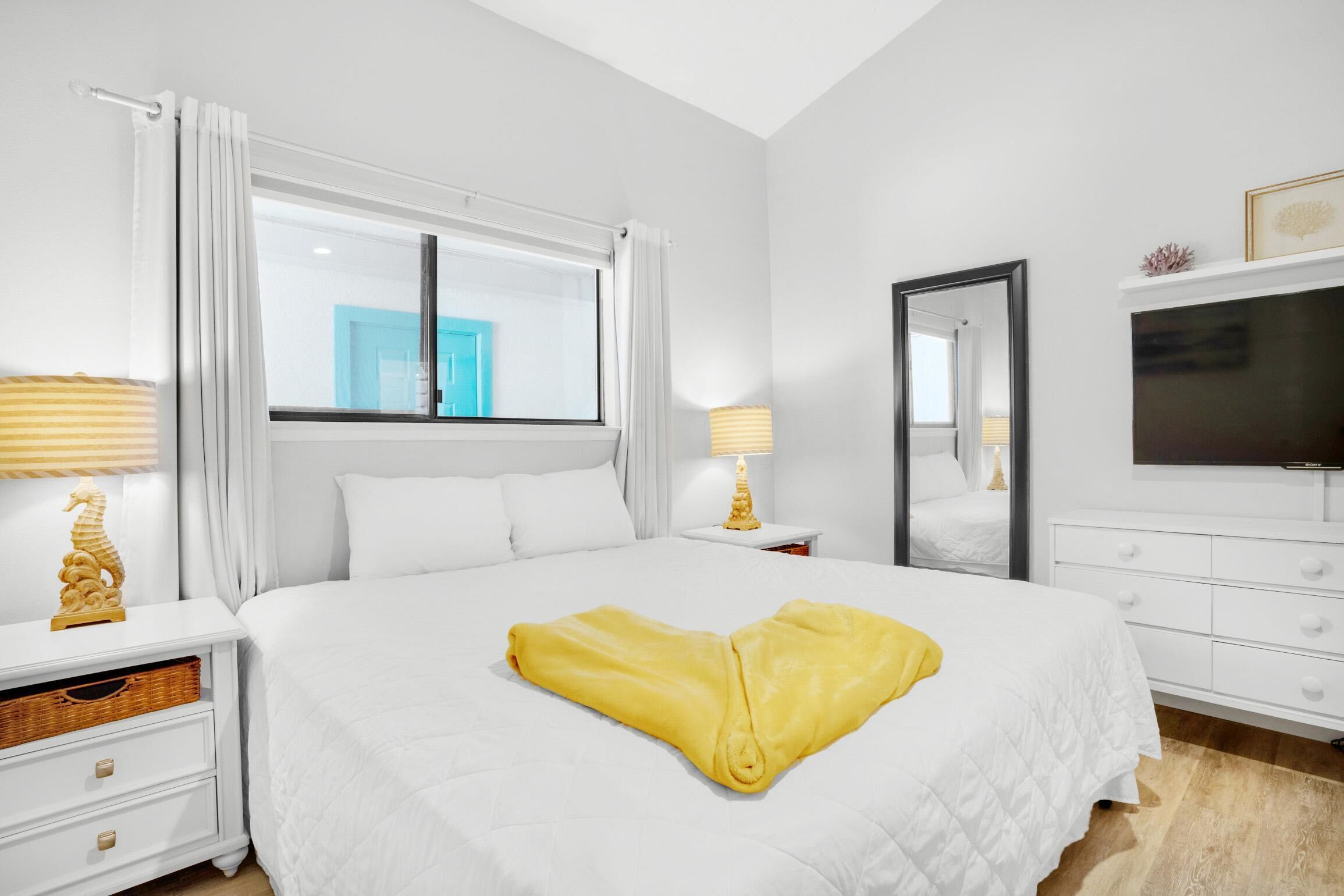 A hotel room with a large bed, white bedding, a flat-screen TV, a bedside table with lamps, and a window with curtains.