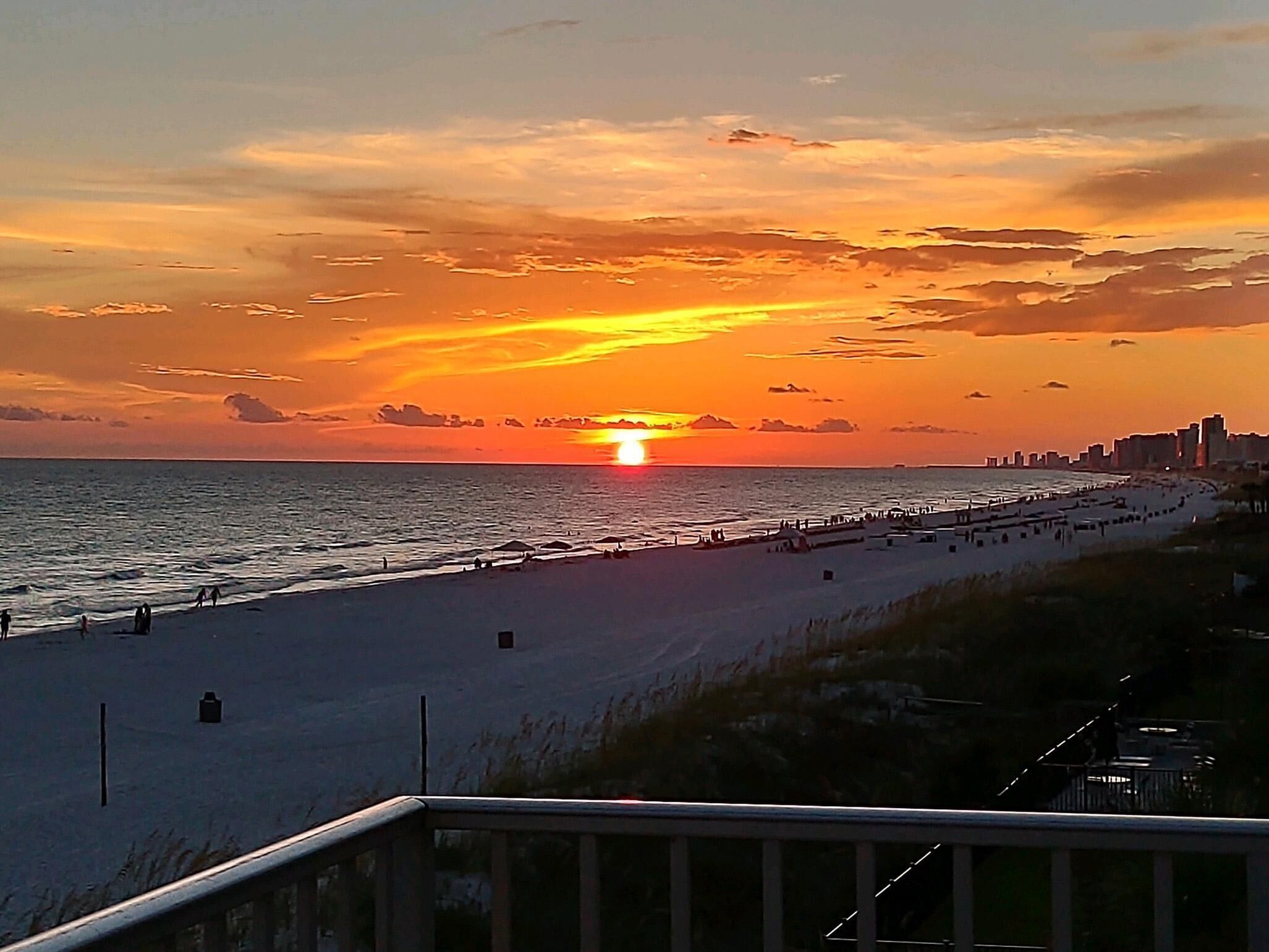 Ein Strand bei Sonnenuntergang, im Hintergrund eine Stadtansicht und im Vordergrund eine Holzbrüstung.