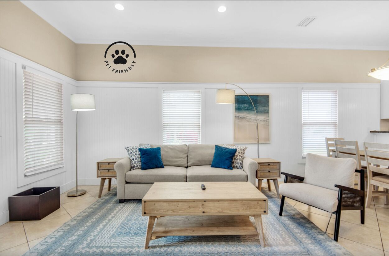 Un salon avec un canapé beige, des coussins bleus, une table basse en bois et un panneau indiquant que les animaux de compagnie sont les bienvenus.
