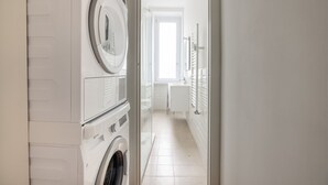 Una sala de lavandería con lavadoras y secadoras apiladas, un baño con inodoro y lavamanos, y un pasillo que conduce a una habitación luminosa con ventana.