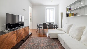Una sala de estar moderna con un televisor de pantalla plana, un mueble de madera para entretenimiento, un sofá blanco, una mesa de comedor con sillas y una estantería.