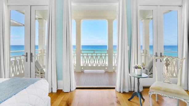 Ein Zimmer mit einem Bett, einem Sessel, einem kleinen Tisch mit Blumen und Meerblick durch offene Türen.