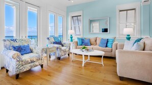 Ein geräumiges Wohnzimmer mit hellblauer Wand, großen Fenstern und Holzboden. Zwei Sessel, ein Sofa und ein Couchtisch sind vorhanden.