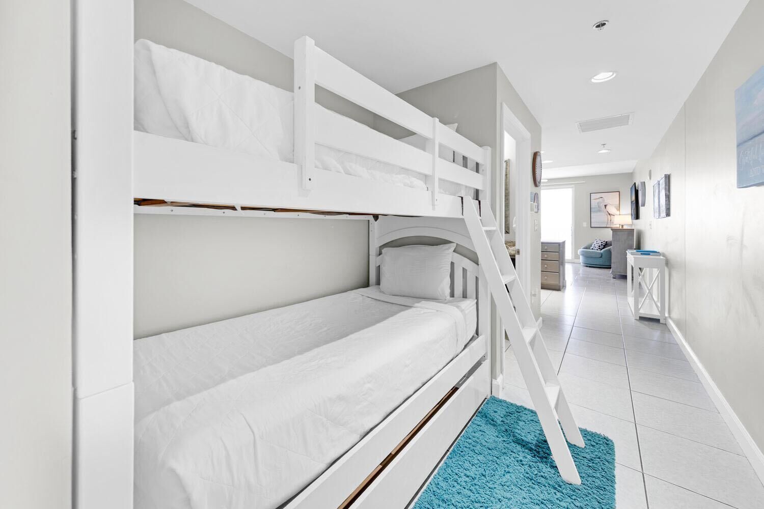 A bunk bed room with a blue rug, white walls, and a hallway leading to a living area.