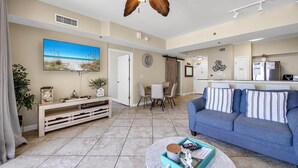 A living room with a blue sofa, a coffee table, a TV, and a dining area with chairs and a table. - Shores of Panama 1212 (Panama City Beach)