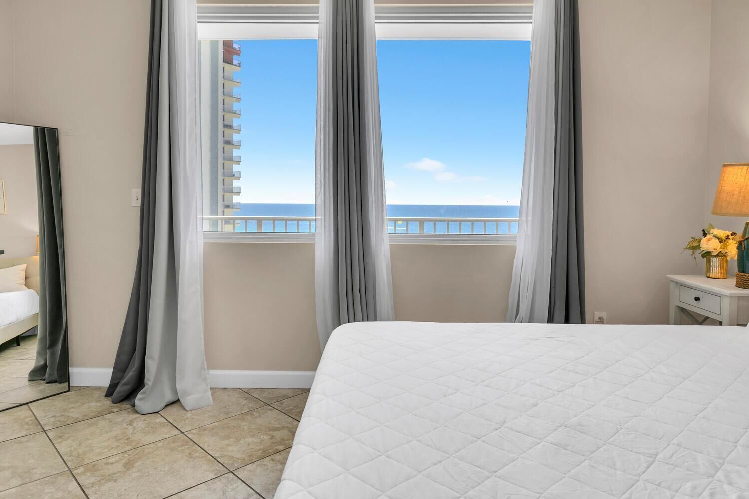 A hotel room with a bed, a nightstand, a lamp, and a view of the ocean through large windows.