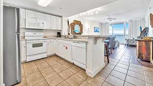 A modern kitchen with white cabinets, a microwave, stove, and dishwasher. There is a large mirror above the sink and a view of the balcony in the background.