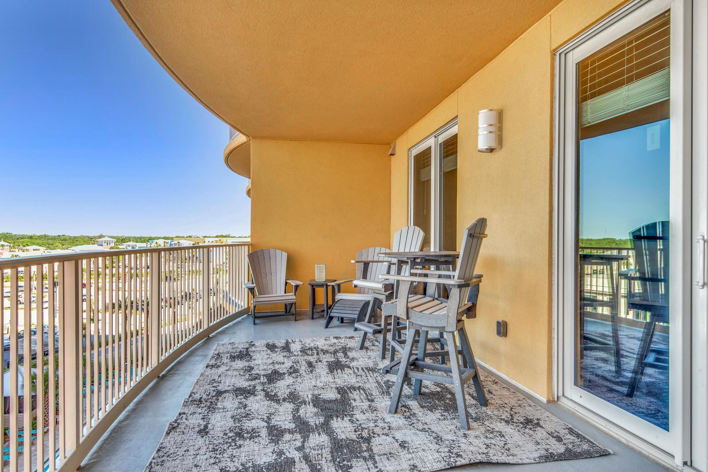 A balcony with a view of a parking lot and buildings, featuring a table and chairs, a rug, and a sliding glass door.