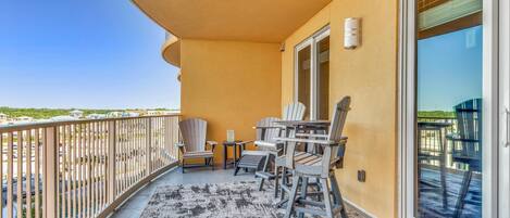 A balcony with a view of a parking lot and buildings, featuring a table and chairs, a rug, and a sliding glass door.