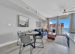A modern living room with a round dining table, grey chairs, a blue sofa, and a balcony with a view of buildings and the sea.