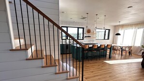 Een ruime woonkamer met een houten trap, een eettafel met stoelen, en een keuken met uitzicht op een bos.