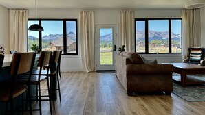 Een ruime woonkamer met houten vloer, een bruine bank, een eettafel met stoelen en grote ramen met uitzicht op de bergen.