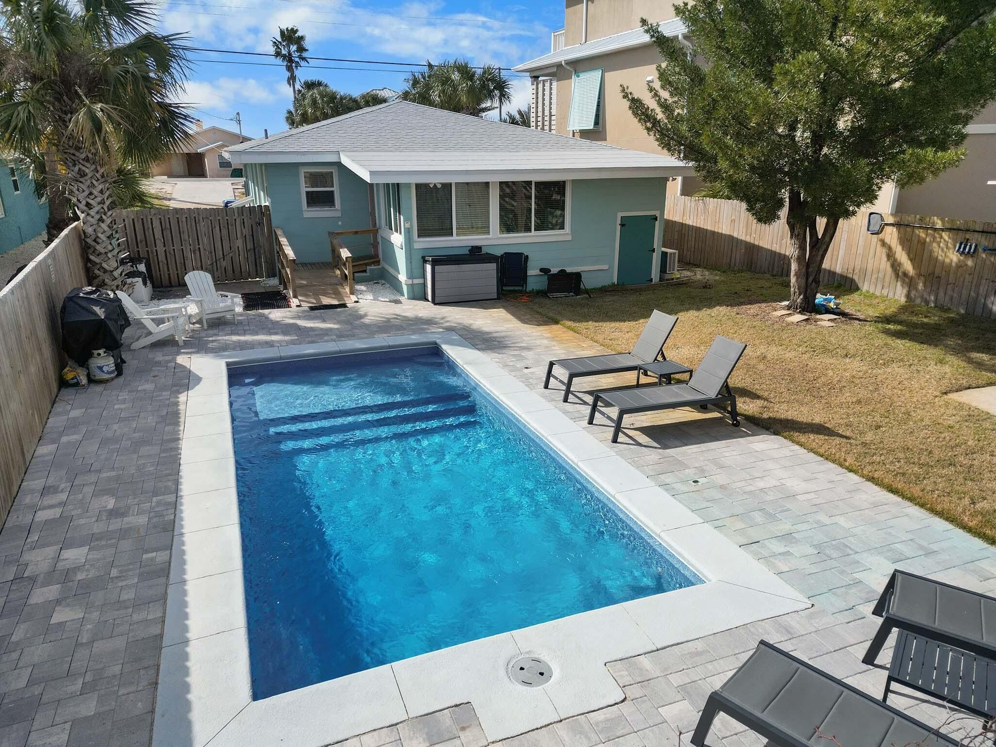 A rectangular swimming pool with a tiled deck, surrounded by lounge chairs and a small house in the background.