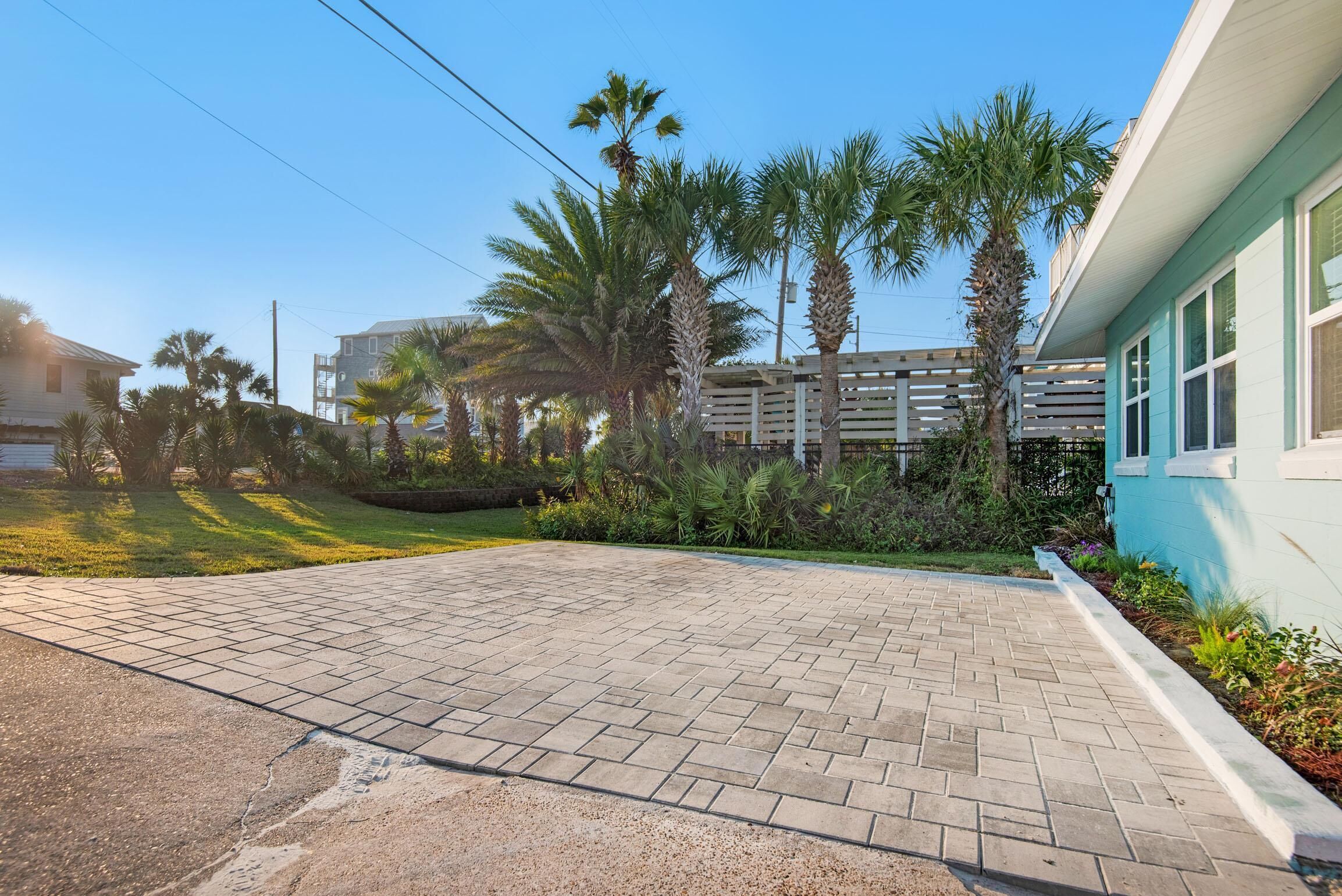 A paved driveway leading to a building with a light blue exterior, surrounded by palm trees and a wooden fence.