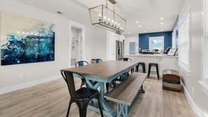 A dining area with a wooden table and benches, a large abstract painting, and a kitchen with blue tiles. - Emerald Flamingo (Panama City Beach)