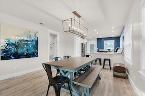 A dining area with a wooden table and benches, a large abstract painting, and a kitchen with blue tiles. - Emerald Flamingo (Panama City Beach)