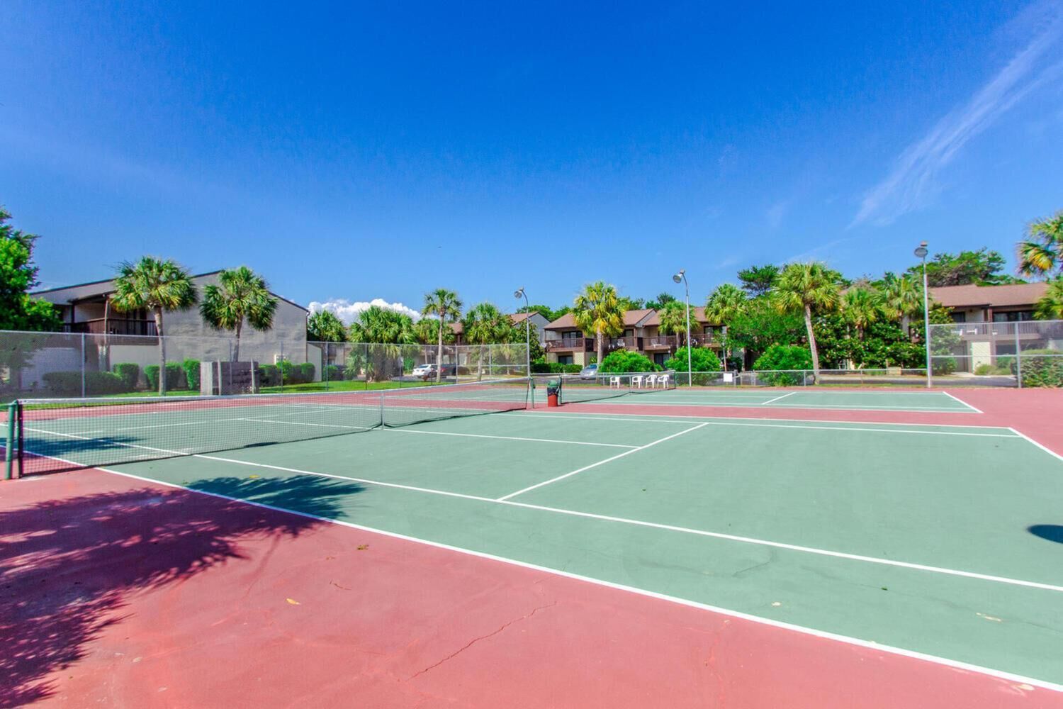 An outdoor tennis court with a net, surrounded by a fence and palm trees.